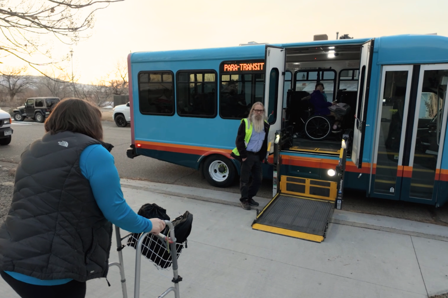 A person using a walker goes in front of a Grand Valley Transit bus with a wheelchair ramp lowered to the sidewalk. A bus driver wearing a green safety vest stands by the doorway.